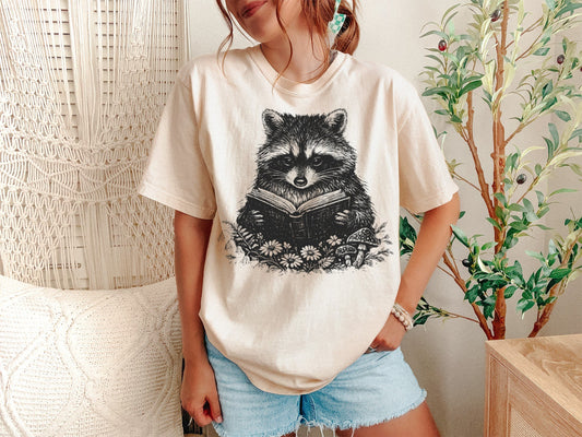 Person wearing a white t-shirt with a raccoon design, standing in a room with a plant and wicker chair.