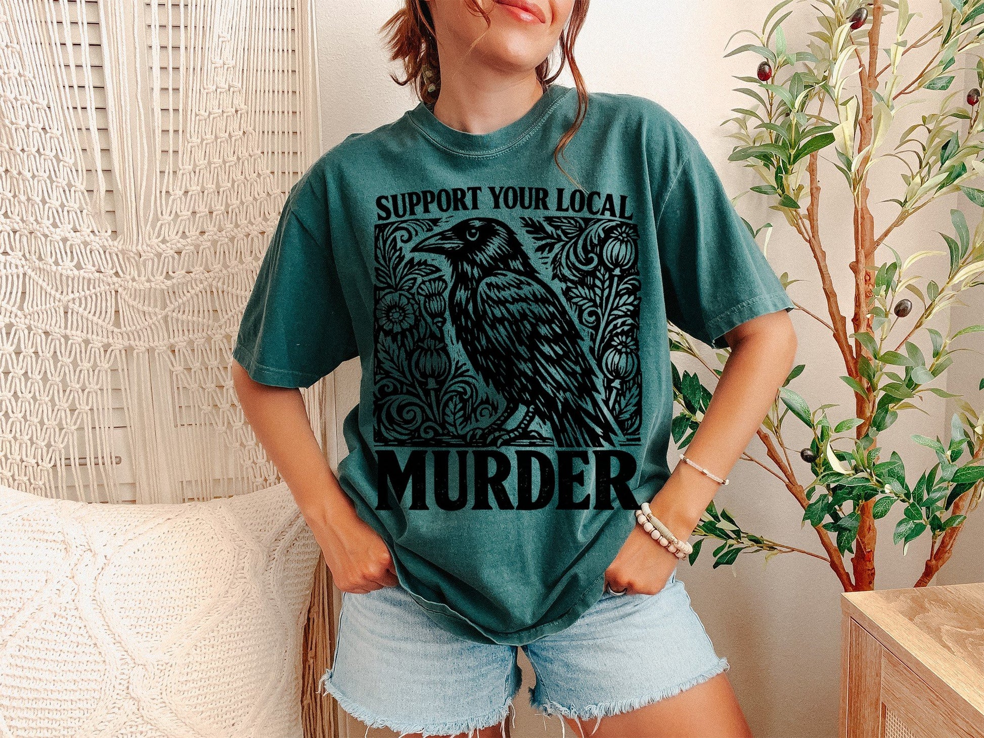 A Blue Spruce Unisex Garment-Dyed T-shirt mockup (front view) showing the support your local murder crow shirt - funny bird lover gift tee print on the front. The artwork highlights crow shirt, murder of crows, support local murder, bird lover gift, crowcore aesthetic, goblincore shirt, funny bird pun, dark academia shirt, witchy crow tee, raven shirt, describing the graphic and theme on the garment.