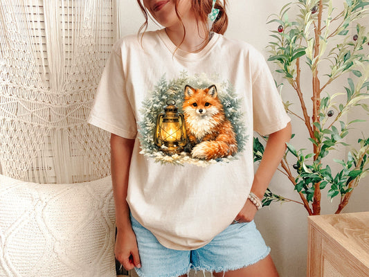 A Ivory Unisex Garment-Dyed T-shirt mockup (front view) showing the cottagecore winter fox shirt - vintage woodland aesthetic tee print on the front. The artwork highlights woodland aesthetic, forestcore clothing, fox lover gift, vintage fox tee, embroidered fox, winter aesthetic, nature lover gift, scandinavian art, cozy winter top, dark academia fox, describing the graphic and theme on the garment.