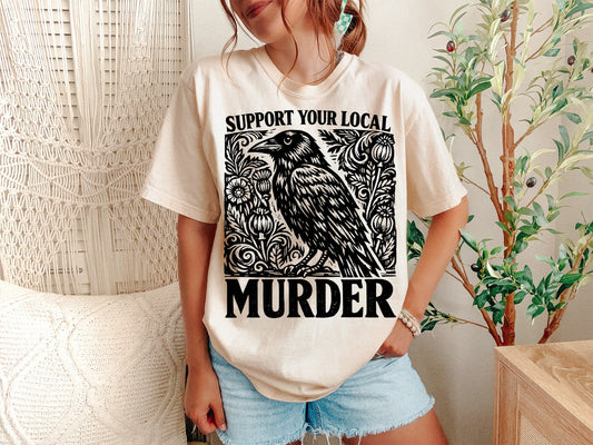 A Ivory Unisex Garment-Dyed T-shirt mockup (front view) showing the support your local murder crow shirt - funny bird lover gift tee print on the front. The artwork highlights support local murder, bird lover gift, crowcore aesthetic, goblincore shirt, funny bird pun, dark academia shirt, witchy crow tee, raven shirt, vintage crow shirt, nature lover gift, describing the graphic and theme on the garment.