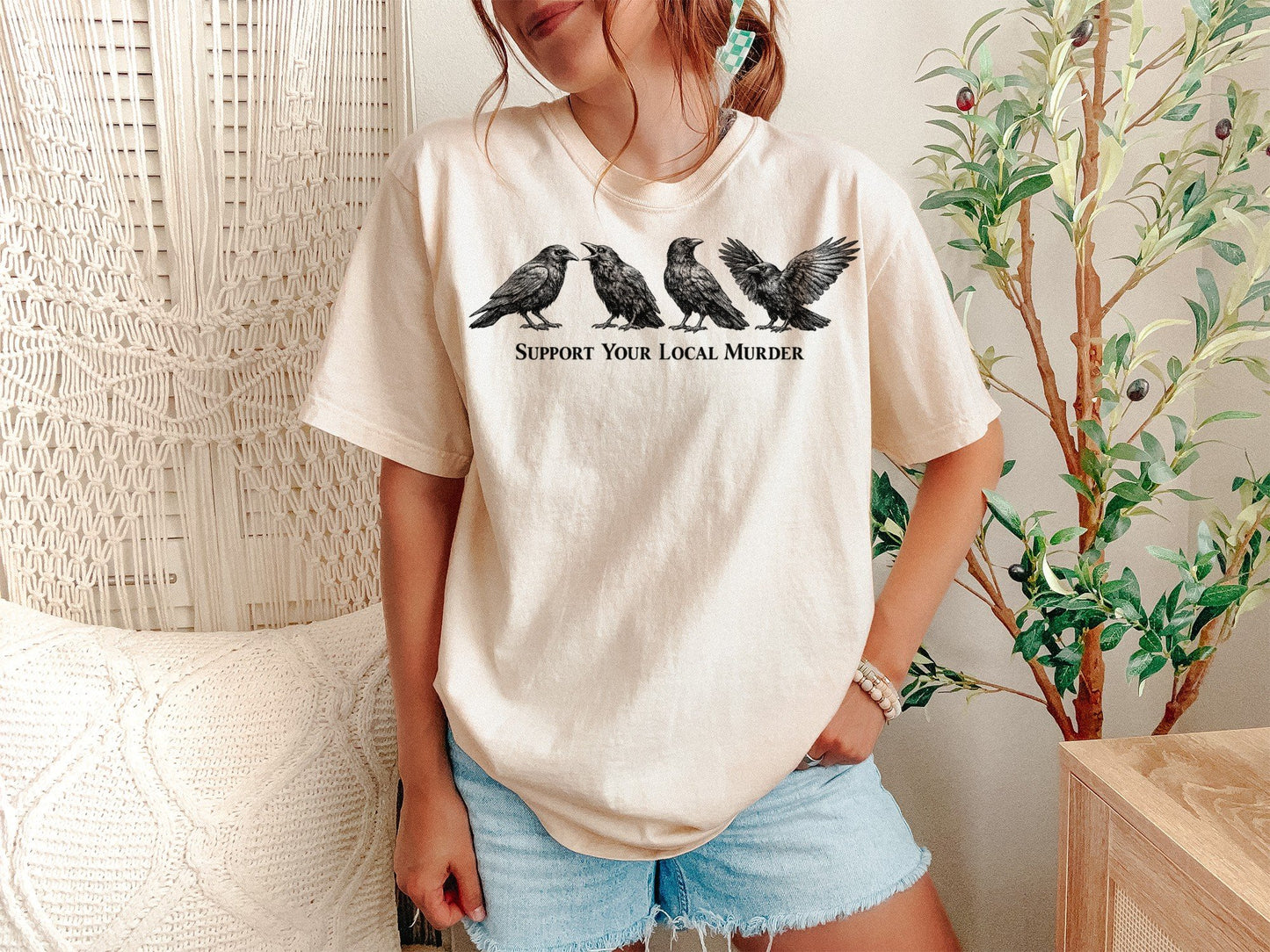 A Ivory Unisex Garment-Dyed T-shirt mockup (front view) showing the support your local murder crow shirt - funny bird lover gift - dark academia raven tee print on the front. The artwork highlights support local murder, funny bird shirt, goblincore, dark academia, raven shirt, bird watcher gift, goth aesthetic, crow lover gift, witchy clothing, cottagecore shirt, describing the graphic and theme on the garment.