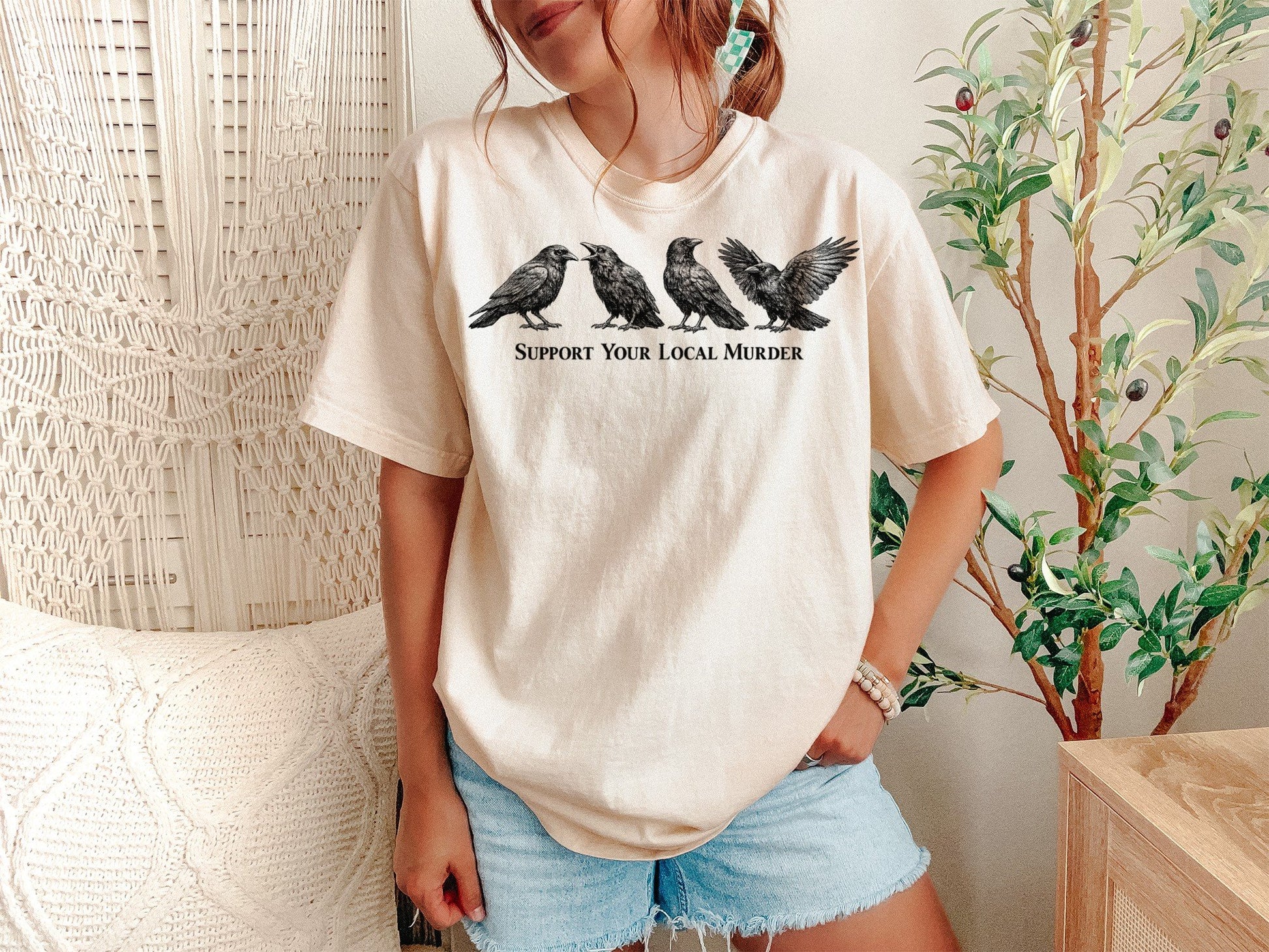 A Ivory Unisex Garment-Dyed T-shirt mockup (front view) showing the support your local murder crow shirt - funny bird lover gift - dark academia raven tee print on the front. The artwork highlights support local murder, funny bird shirt, goblincore, dark academia, raven shirt, bird watcher gift, goth aesthetic, crow lover gift, witchy clothing, cottagecore shirt, describing the graphic and theme on the garment.