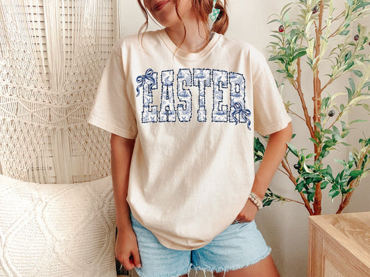 A Ivory Unisex Garment-Dyed T-shirt mockup (front view) showing the easter coquette chinoiserie bunny shirt - blue and white toile bow tee print on the front. The artwork highlights coquette bow shirt, grandmillennial, blue and white, easter bunny shirt, preppy easter, toile bunny, coquette aesthetic, chinoiserie shirt, easter gift for her, spring fashion, describing the graphic and theme on the garment.