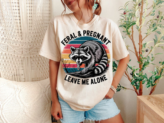 A Ivory Unisex Garment-Dyed T-shirt mockup (front view) showing the feral and pregnant shirt - funny raccoon maternity tee - unhinged pregnancy announcement print on the front. The artwork highlights funny maternity, pregnancy humor, unhinged mom, feral mom era, trash panda, dont touch me, pregnancy reveal, baby announcement, mom to be gift, comfort colors, describing the graphic and theme on the garment.