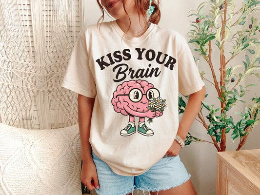 A Ivory Unisex Garment-Dyed T-shirt mockup (front view) showing the retro kiss your brain shirt - groovy kindergarten teacher tee print on the front. The artwork highlights kindergarten tee, preschool teacher, retro teacher shirt, groovy teacher, teacher gift, back to school, mental health shirt, sped teacher shirt, elementary teacher, teacher squad, describing the graphic and theme on the garment.