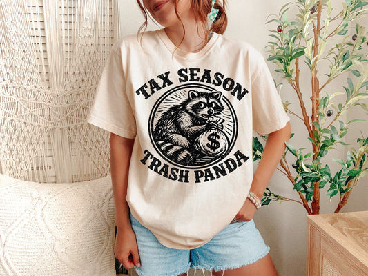 A Ivory Unisex Garment-Dyed T-shirt mockup (front view) showing the funny tax season trash panda shirt - raccoon accountant tee print on the front. The artwork highlights funny raccoon tee, accountant gift, cpa shirt, tax professional, raccoon meme, funny tax shirt, dad tax shirt, bookkeeper gift, tax season 2026, trash panda gift, describing the graphic and theme on the garment.