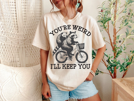 A Ivory Unisex Garment-Dyed T-shirt mockup (front view) showing the youre weird ill keep you shirt - funny raccoon and opossum couple tee print on the front. The artwork highlights funny raccoon tee, trash panda shirt, opossum shirt, matching couple, best friend gift, sarcastic shirt, weird couple gift, raccoon hug tee, anniversary gift, quirky t-shirt, describing the graphic and theme on the garment.