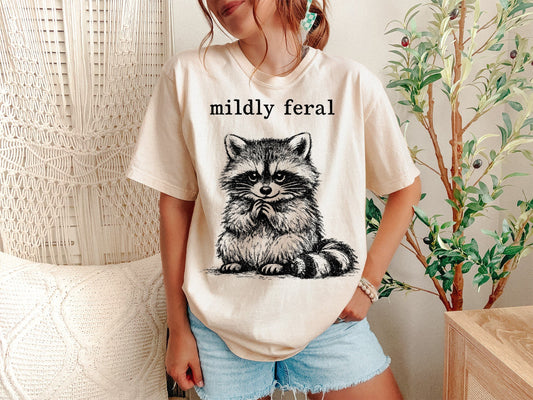 Vintage-style black and white ink illustration of a fluffy raccoon sitting with its paws tucked under its chin. The text "mildly feral" is written in a typewriter-style serif font above the raccoon.