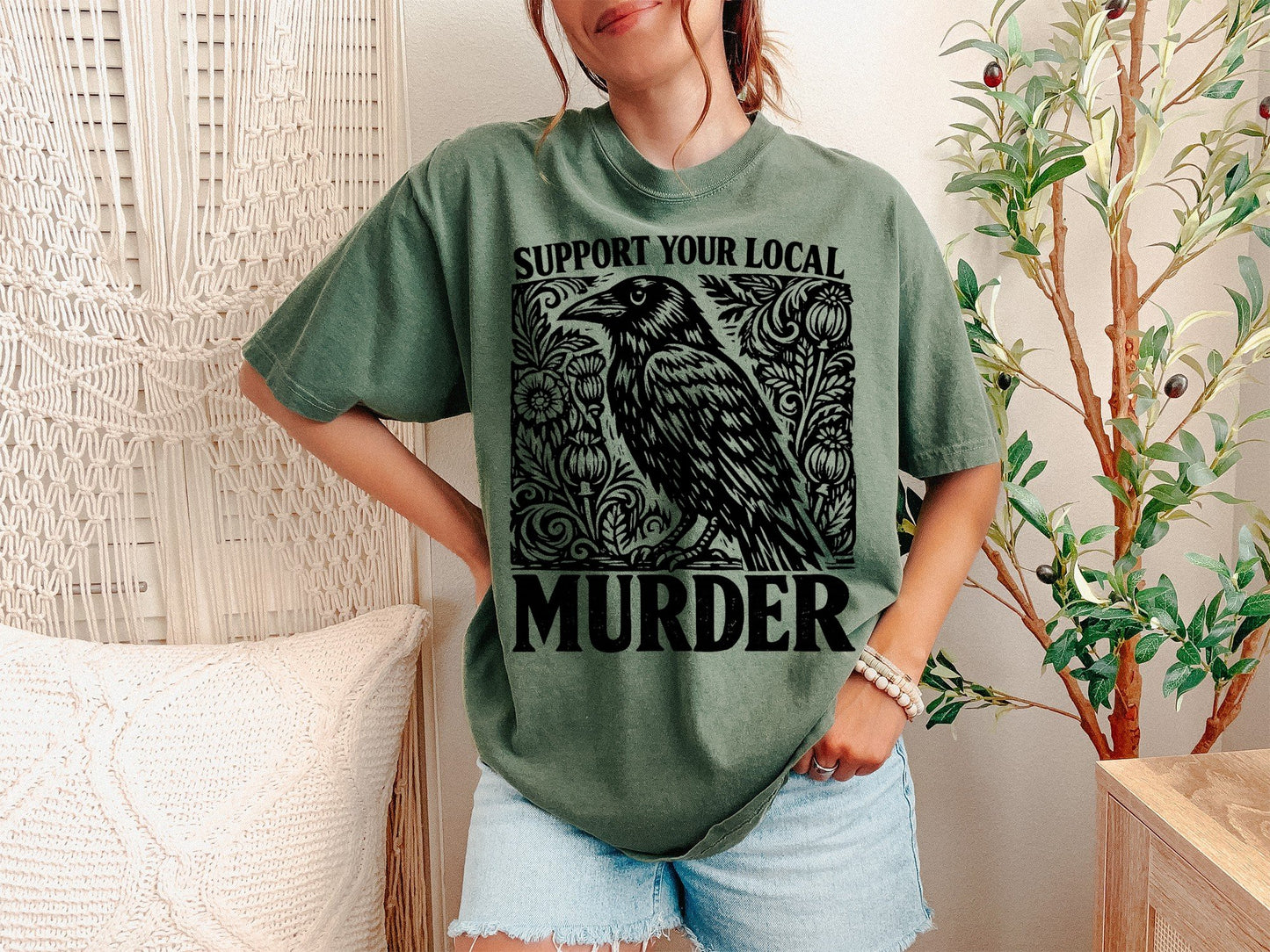 A Moss Unisex Garment-Dyed T-shirt mockup (front view) showing the support your local murder crow shirt - funny bird lover gift tee print on the front. The artwork highlights murder of crows, support local murder, bird lover gift, crowcore aesthetic, goblincore shirt, funny bird pun, dark academia shirt, witchy crow tee, raven shirt, vintage crow shirt, describing the graphic and theme on the garment.