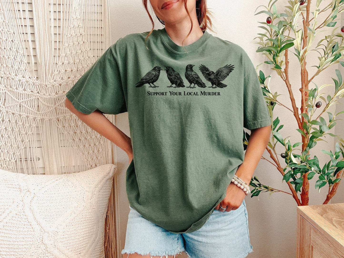 A Moss Unisex Garment-Dyed T-shirt mockup (front view) showing the support your local murder crow shirt - funny bird lover gift - dark academia raven tee print on the front. The artwork highlights garment-dyed, t-shirt, crow shirt, murder of crows, support local murder, funny bird shirt, goblincore, dark academia, raven shirt, bird watcher gift, describing the graphic and theme on the garment.